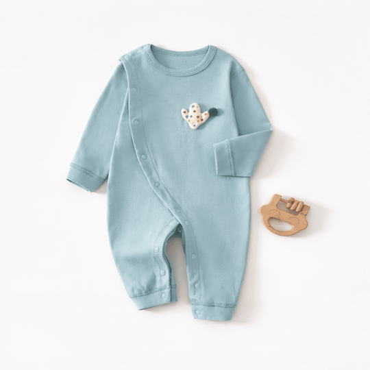 Pyjama pour bébé bleu clair à manches longues avec boutons pression sur le côté et petit motif cactus sur la poitrine, posé à plat sur un fond clair avec un jouet de dentition en bois à côté.