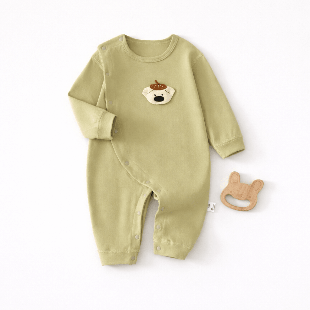 Pyjama pour bébé vert clair à manches longues avec fermeture à boutons pression sur le côté et motif tête de chien sur la poitrine, présenté à plat sur un fond clair avec un jouet de dentition en bois en forme de lapin à côté.