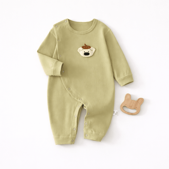 Pyjama pour bébé vert clair à manches longues avec fermeture à boutons pression sur le côté et motif tête de chien sur la poitrine, présenté à plat sur un fond clair avec un jouet de dentition en bois en forme de lapin à côté.