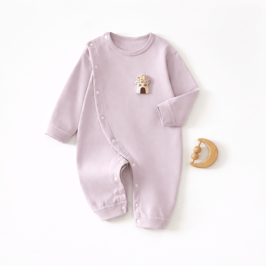 Pyjama pour bébé violet pastel à manches longues avec fermeture à boutons pression sur le côté et motif petite maison sur la poitrine, présenté à plat sur un fond clair avec un jouet de dentition en bois en forme de lune.