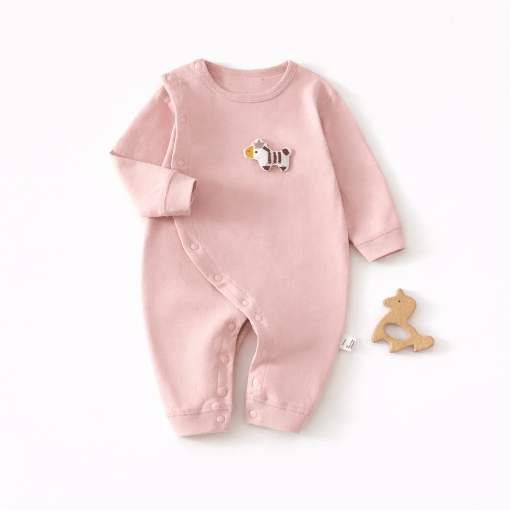 Pyjama pour bébé rose clair à manches longues avec fermeture à boutons pression sur le côté et motif animal sur la poitrine, présenté à plat sur un fond clair avec un jouet de dentition en bois en forme d’animal à côté.