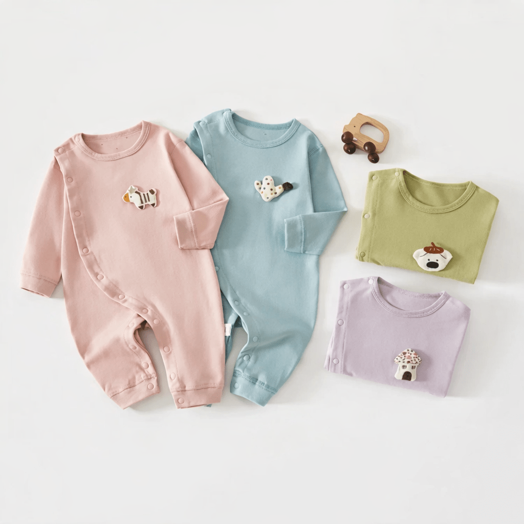 Quatre pyjamas pour bébé en coton, de couleurs pastel (rose, bleu, vert et violet) avec petits motifs d’animaux, disposés à plat sur un fond clair, accompagnés d’un jouet en bois.