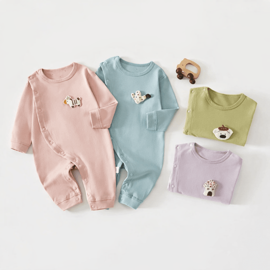 Quatre pyjamas pour bébé en coton, de couleurs pastel (rose, bleu, vert et violet) avec petits motifs d’animaux, disposés à plat sur un fond clair, accompagnés d’un jouet en bois.