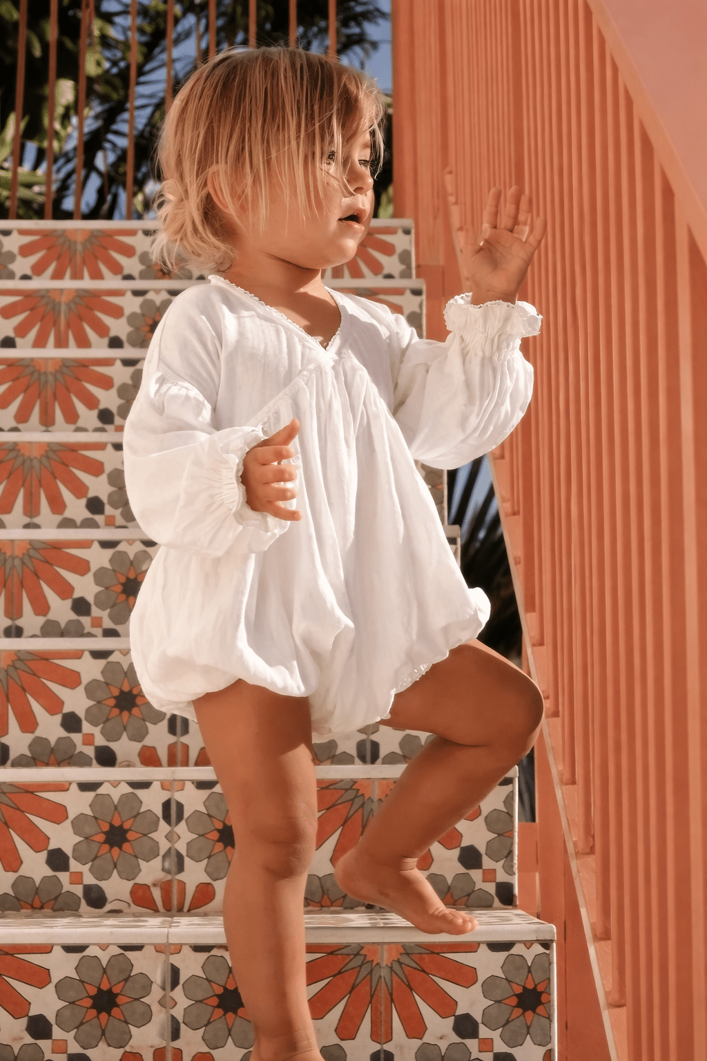 Bébé fille portant une barboteuse blanche en coton avec manches longues, debout sur un escalier coloré au soleil.