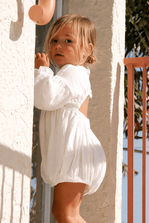Petite fille portant une barboteuse blanche en coton avec manches longues, debout près d’un mur au soleil, tenue bébé élégante été.