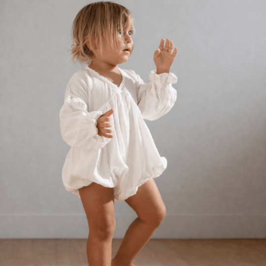 Petite fille portant une barboteuse blanche en coton avec manches longues, debout pieds nus dans un intérieur lumineux, tenue bébé élégante.