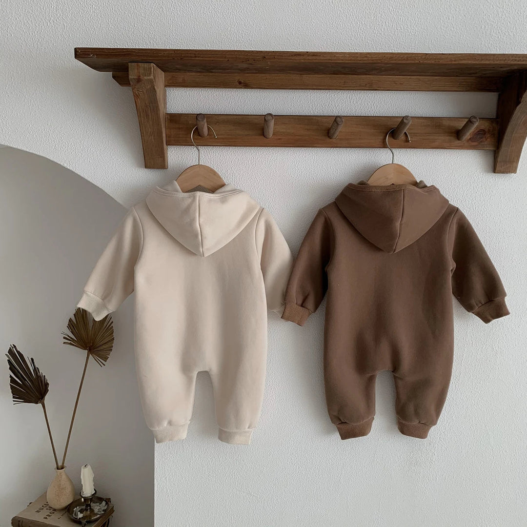 Dos des combinaisons bébé beige et marron à capuche, suspendues sur une étagère murale.