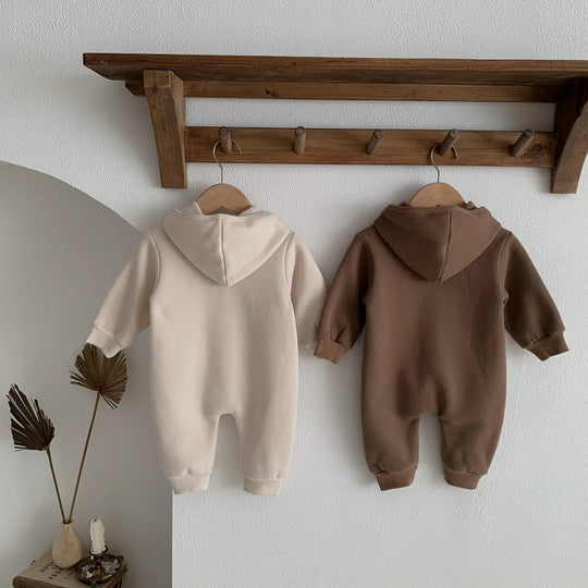 Dos des combinaisons bébé beige et marron à capuche, suspendues sur une étagère murale.