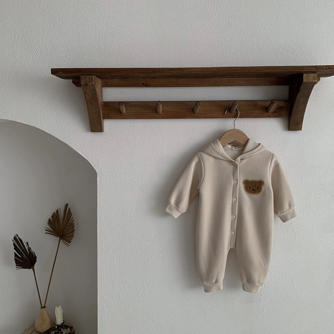 Combinaison bébé beige à capuche avec patch ourson marron, suspendue sur un cintre.