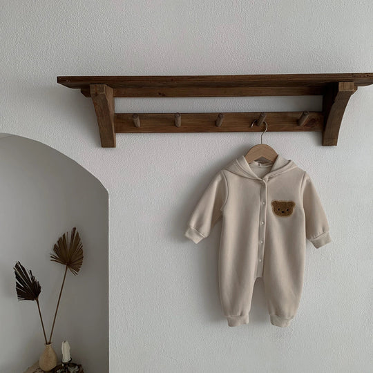 Combinaison bébé beige à capuche avec patch ourson marron, suspendue sur un cintre.