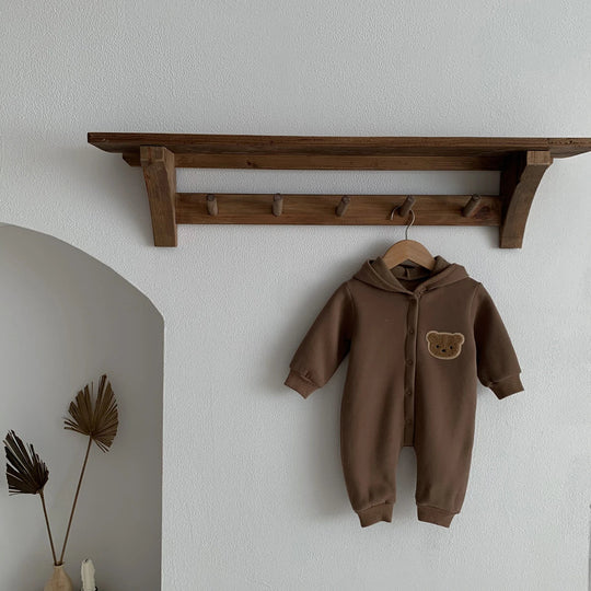 Combinaison bébé marron à capuche avec motif ourson, suspendue sur un cintre sous une étagère en bois.