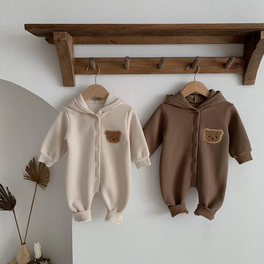 Deux combinaisons bébé beige et marron avec capuche et ourson, suspendues à une étagère décorative.