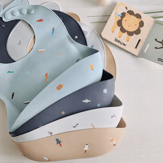 Ensemble de bavoirs bébé en silicone empilés, couleurs pastel (bleu, gris, beige), avec motifs ludiques, posés sur une table avec un livre enfant
