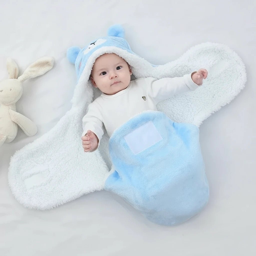 Bébé emmailloté dans une couverture bleue à capuche tête d’ours Kalinatur, posé sur un drap blanc avec un doudou lapin à côté.