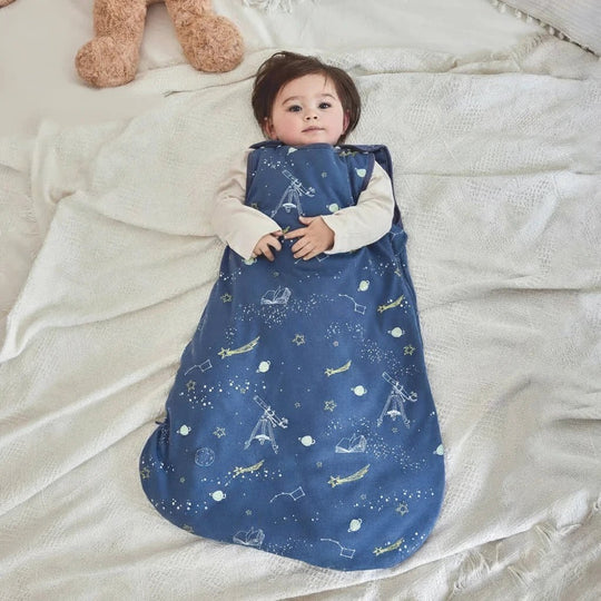 Bébé allongé portant une gigoteuse bleu nuit en coton à motifs d’étoiles et de fusées, collection Kalinatur.
