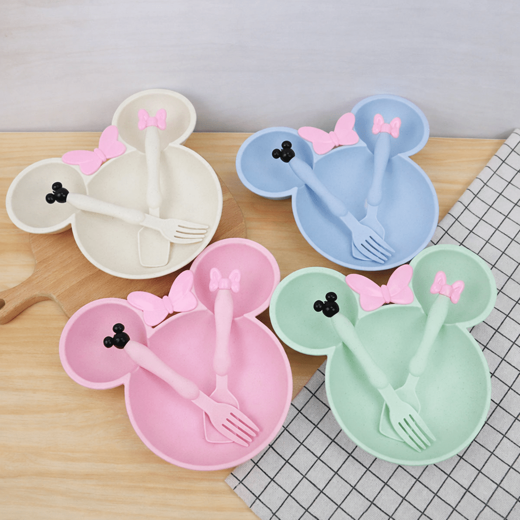 Quatre assiettes pour enfants en forme de tête de souris, de couleurs pastel crème, bleu, rose et vert, sont posées sur une table en bois. Chaque assiette contient des couverts assortis avec de petits nœuds roses, sur fond de mur clair et tissu quadrillé noir et blanc.