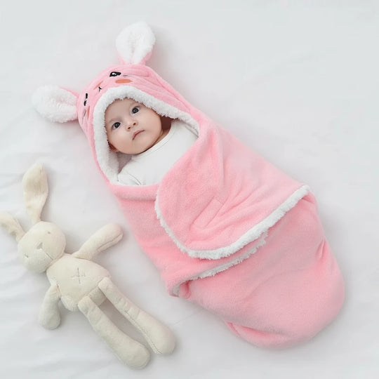 Bébé emmailloté dans une couverture rose douce en forme de lapin avec capuche à oreilles blanches, allongé à côté d’un doudou lapin, marque Kalinatur.

