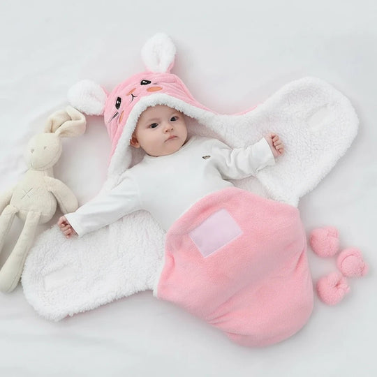 Bébé emmailloté dans une couverture rose en forme de lapin avec capuche à oreilles et intérieur en polaire blanche, à côté d’un doudou lapin, marque Kalinatur.
