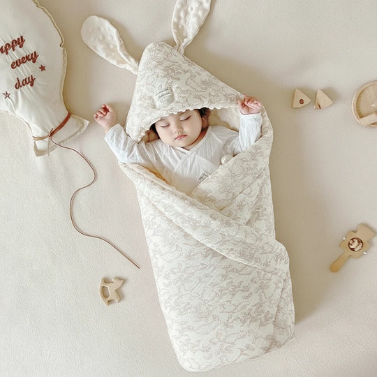 Bébé endormi enveloppé dans une couverture en mousseline de coton avec capuche à oreilles, couleur ivoire, accessoire Kalinatur posé dans un décor doux et naturel.