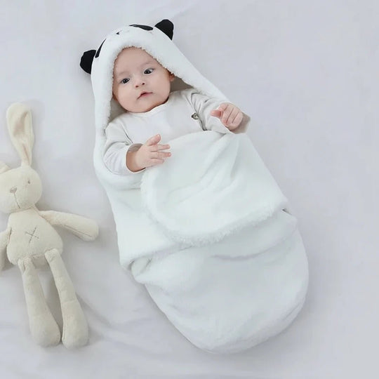 Bébé emmailloté dans une couverture panda blanche Kalinatur, douce et chaude avec capuche à oreilles, allongé sur un drap clair à côté d’un doudou lapin.
