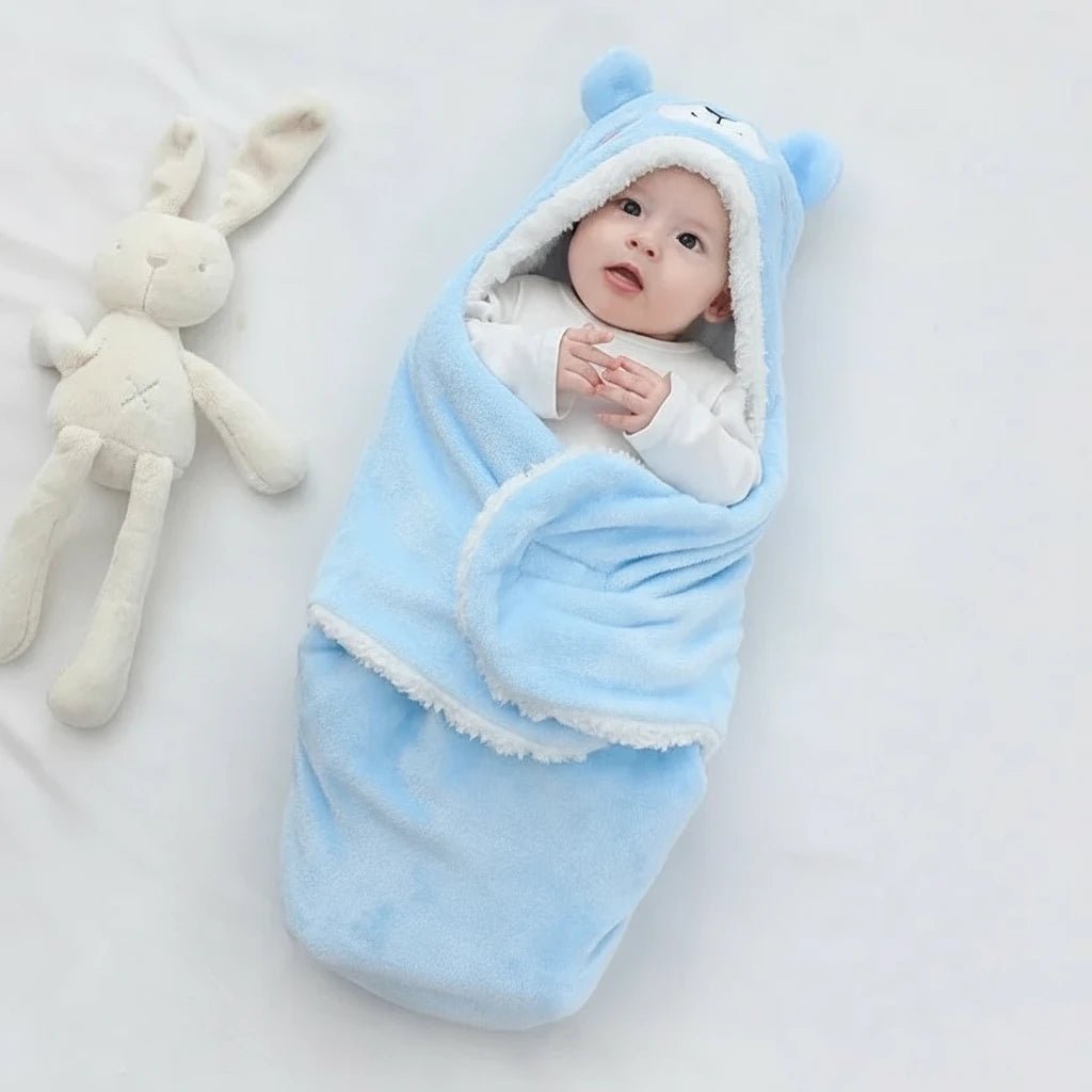 Bébé emmailloté dans une couverture d’hiver bleue Kalinatur en forme d’ours, en polaire douce et chaude avec capuche à oreilles, posé sur un drap blanc à côté d’un doudou lapin en peluche.