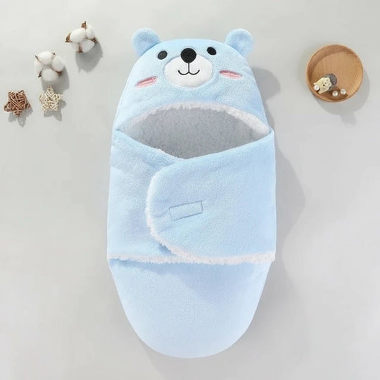 Couverture d’emmaillotage bleue pour bébé Kalinatur, avec capuche tête d’ours et intérieur en polaire blanche douce, posée sur une surface claire avec décor naturel.
