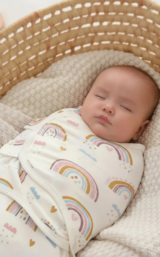 Bébé endormi dans une gigoteuse Kalinatur motif arc-en-ciel, allongé dans un panier en osier sur une couverture beige.