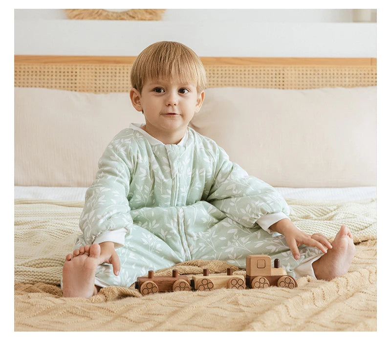 Bébé assis sur un lit portant une gigoteuse à jambes Kalinatur vert doux imprimée de petites branches blanches, jouant avec un train en bois naturel.
