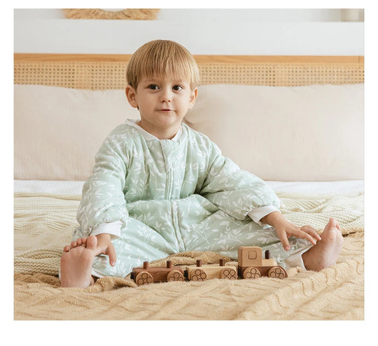 Bébé assis sur un lit portant une gigoteuse à jambes Kalinatur vert doux imprimée de petites branches blanches, jouant avec un train en bois naturel.
