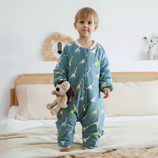 Enfant portant la gigoteuse à jambes Kalinatur motif dinosaures, en coton bio bleu canard, idéale pour l’hiver, debout sur un lit avec une peluche à la main.
