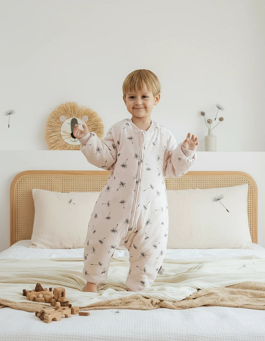 Enfant portant la gigoteuse à jambes Kalinatur rose pâle avec imprimé pissenlits, en coton bio, sur un lit beige dans une chambre douce et naturelle.