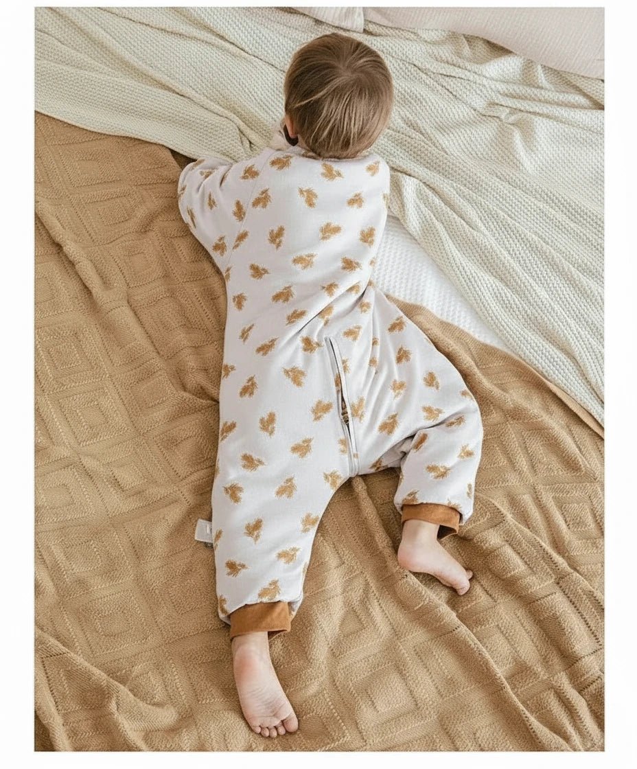 Enfant allongé sur le ventre portant une gigoteuse à jambes Kalinatur en coton bio, imprimée de petites feuilles automnales marron, sur un lit couvert d’une couverture beige et d’un plaid crème.