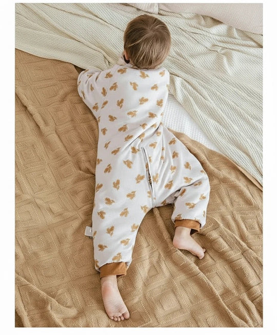 Enfant allongé sur le ventre portant une gigoteuse à jambes Kalinatur en coton bio, imprimée de petites feuilles automnales marron, sur un lit couvert d’une couverture beige et d’un plaid crème.