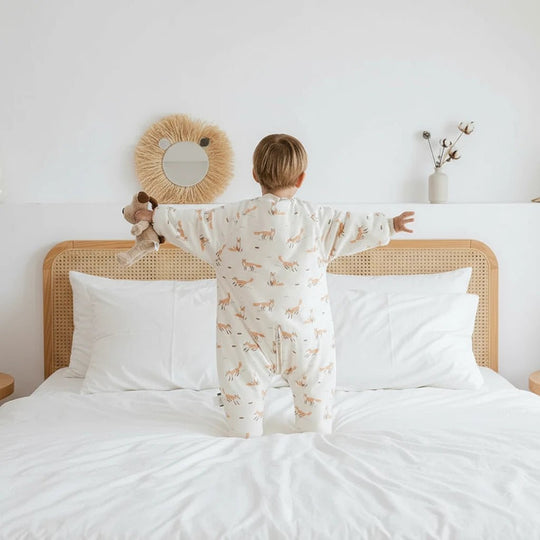Enfant vu de dos portant une gigoteuse à jambes Kalinatur en coton bio, motif renard sur fond beige, debout sur un lit blanc dans une chambre au style naturel et épuré.
