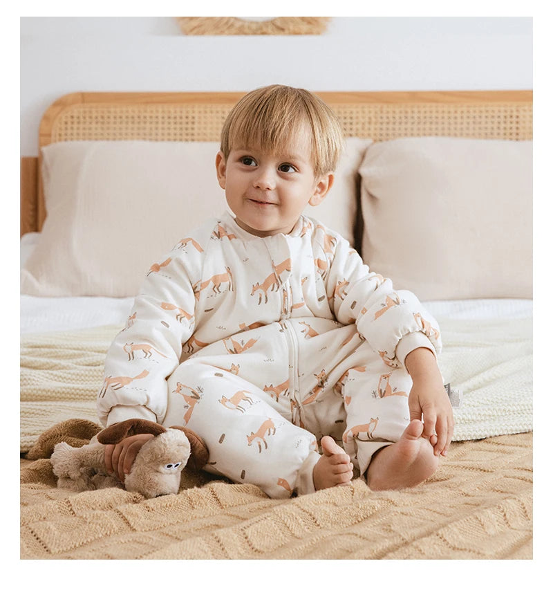Bébé assis sur un lit tenant une peluche, portant la gigoteuse à jambes Kalinatur écrue imprimée de renards, en coton doux, pour un sommeil confortable et sécurisé.
