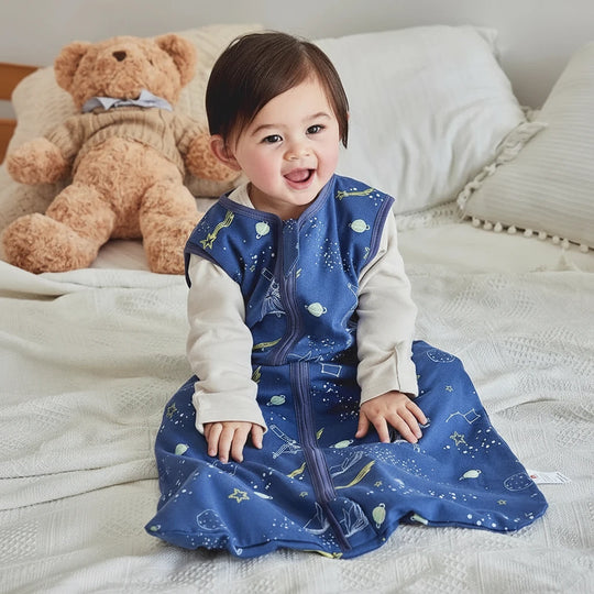 Bébé souriant assis sur un lit, portant une gigoteuse bleue Kalinatur décorée d’étoiles, de planètes et de constellations, offrant chaleur et confort pour les nuits paisibles.
