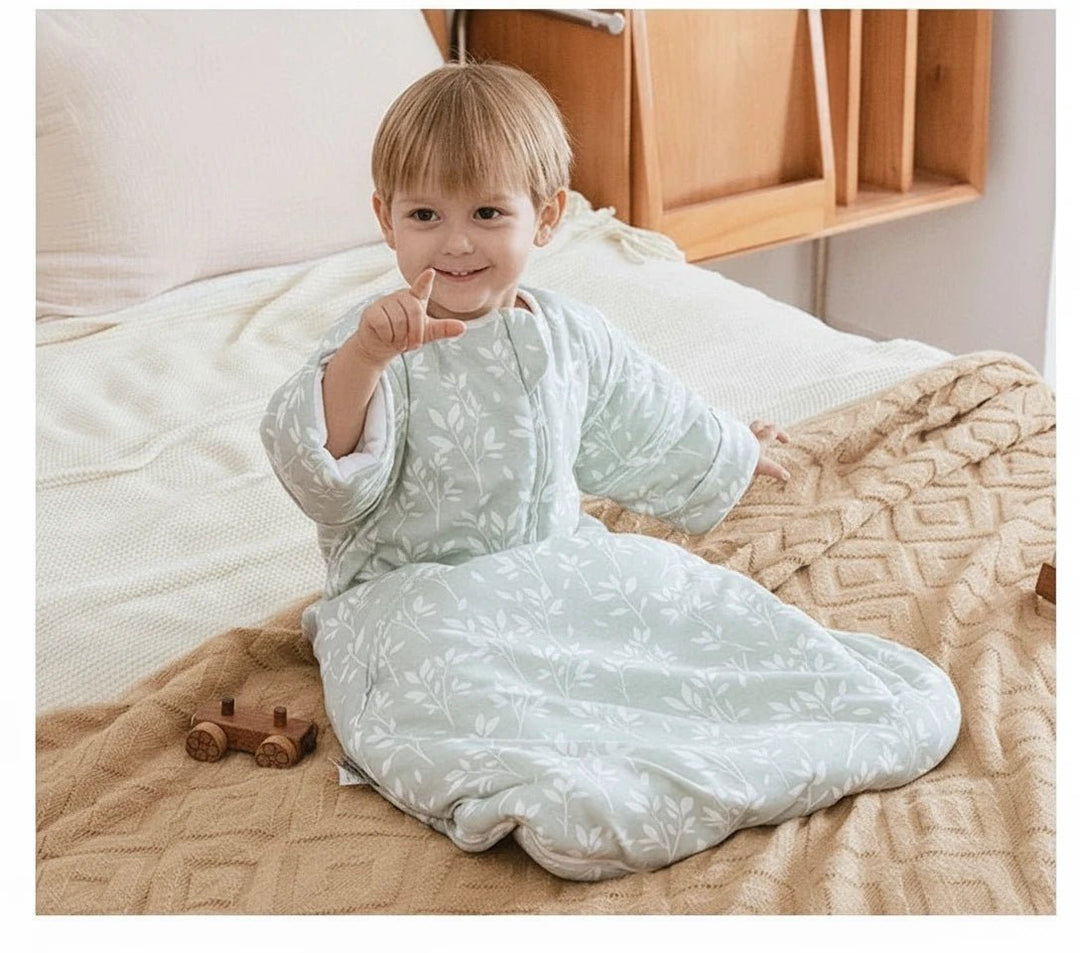 Bébé portant une gigoteuse d’hiver vert sauge Kalinatur à motif feuillage blanc, assis sur un lit beige, souriant et jouant.

