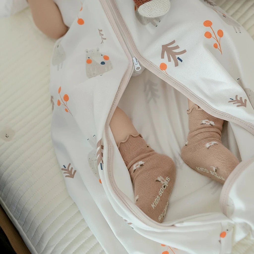 Pieds de bébé vêtus de chaussettes beiges, reposant dans une gigoteuse crème à motifs d’ours et de feuilles orangées, offrant confort et douceur pour les nuits d’automne.
