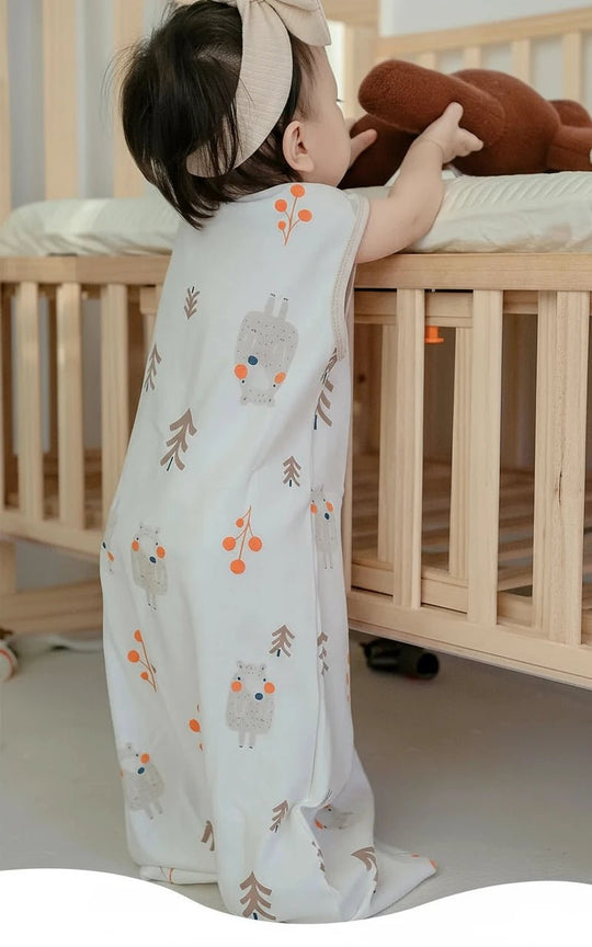 Bébé debout près de son lit en bois, vêtu d’une gigoteuse crème à motifs d’ours et de branches, dans une chambre douce et naturelle, ambiance cocooning.
