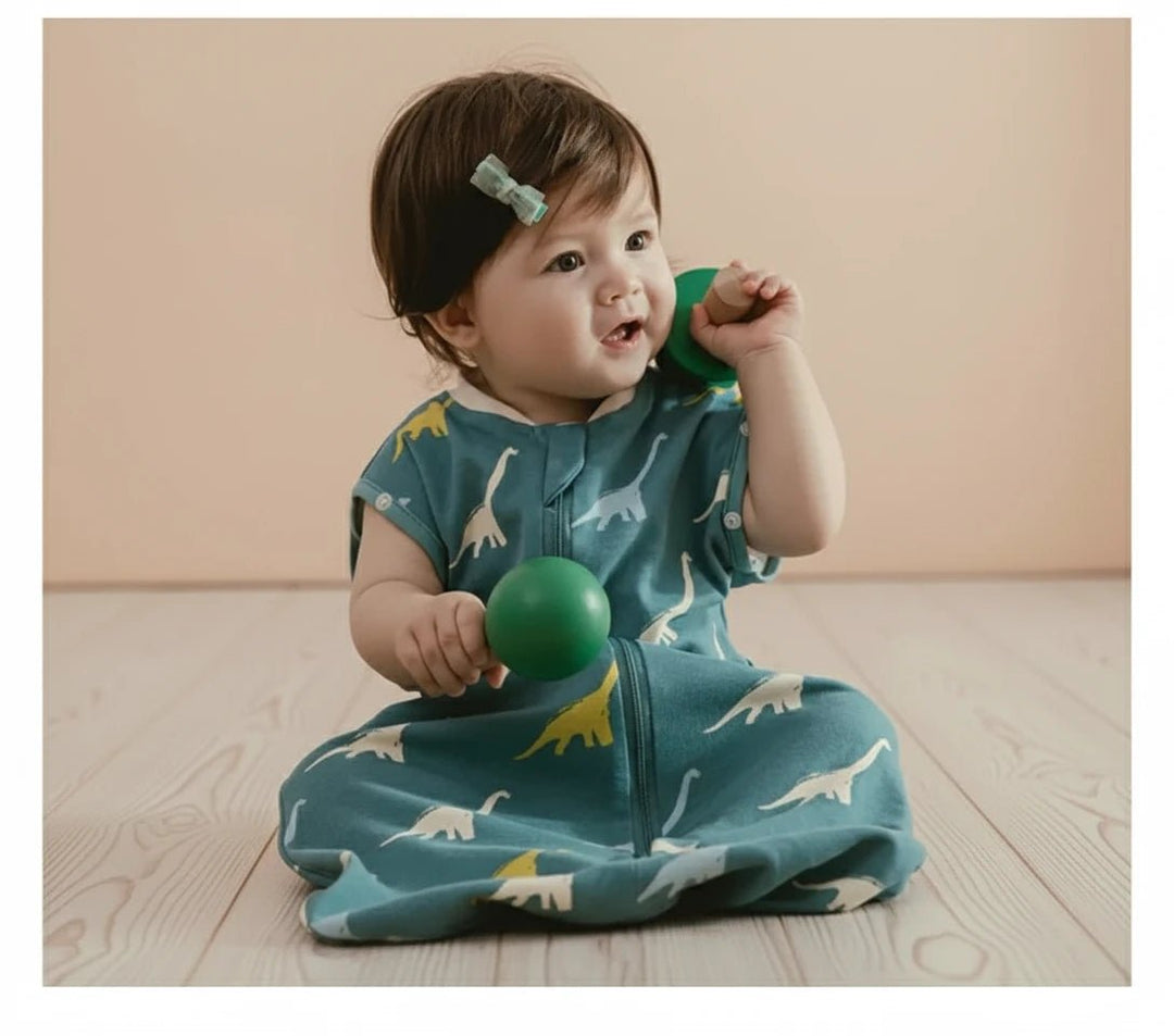 Bébé assis sur un parquet clair portant la gigoteuse d’été Kalinatur bleue à motifs dinosaures, en coton léger et respirant, parfait pour les nuits chaudes.
