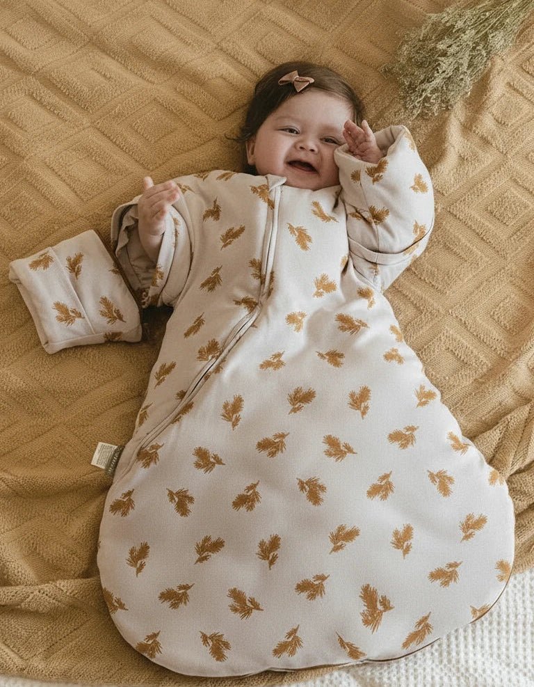 Bébé souriant allongé sur un plaid ocre, portant une gigoteuse d’hiver Kalinatur en coton doux, beige avec motif feuilles marron, chaude et confortable.