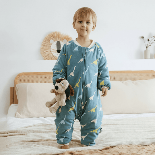 Enfant portant la gigoteuse à jambes Kalinatur motif dinosaures, en coton bio bleu canard, idéale pour l’hiver, debout sur un lit avec une peluche à la main.
