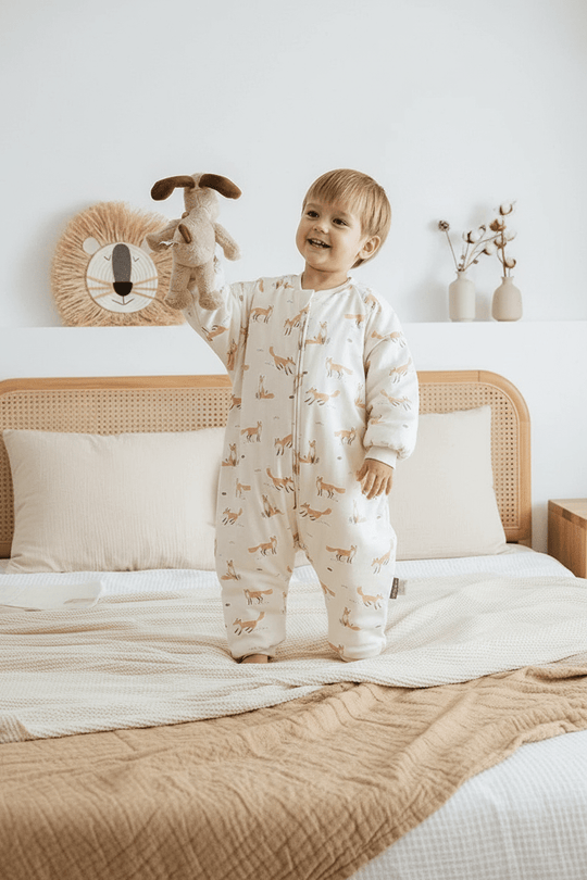 Enfant debout sur un lit portant la gigoteuse à jambes Kalinatur écrue avec imprimé renards, en coton doux, dans une chambre naturelle et chaleureuse.