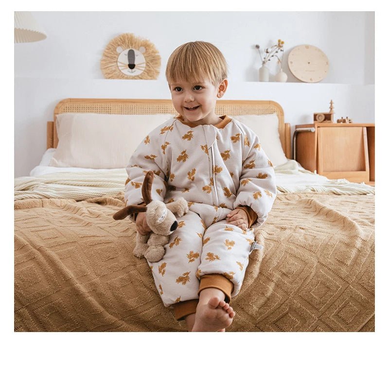 Petit garçon assis sur un lit, tenant une peluche, vêtu d’une gigoteuse à jambes Kalinatur beige à motifs de feuilles marron, dans une chambre douce et naturelle.
