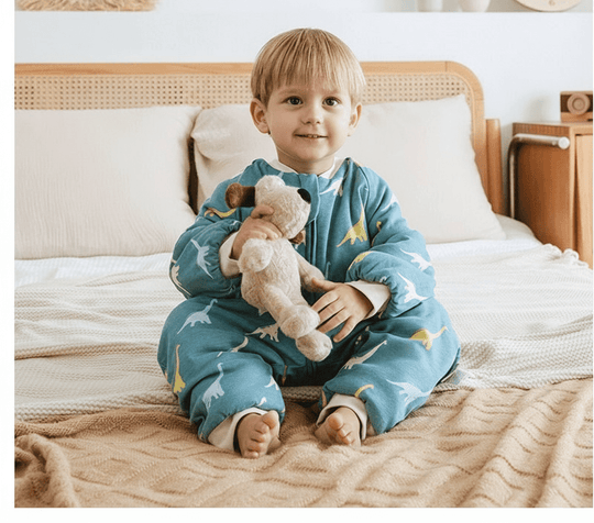 Petit garçon assis sur un lit tenant une peluche, vêtu de la gigoteuse à jambes Kalinatur motif dinosaures bleus et jaunes, en coton bio chaud et respirant pour l’hiver.
