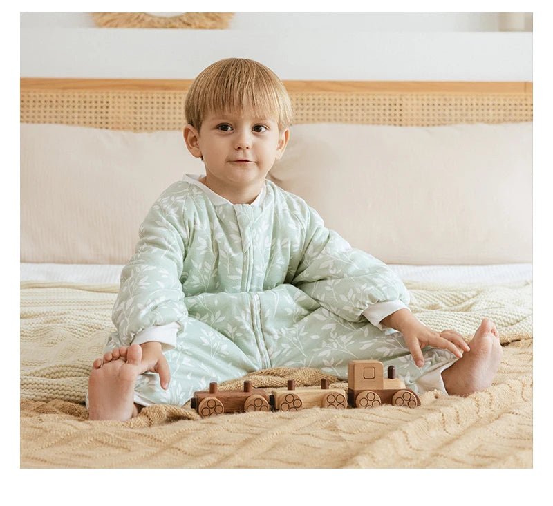 Bébé assis sur un lit portant une gigoteuse à jambes Kalinatur vert doux imprimée de petites branches blanches, jouant avec un train en bois naturel.
