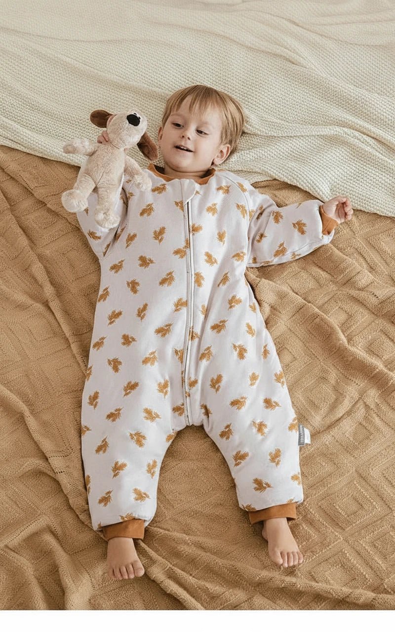 Enfant jouant sur un lit beige, vêtu d’une gigoteuse à jambes Kalinatur motif feuilles automnales, chaude et confortable pour l’hiver.