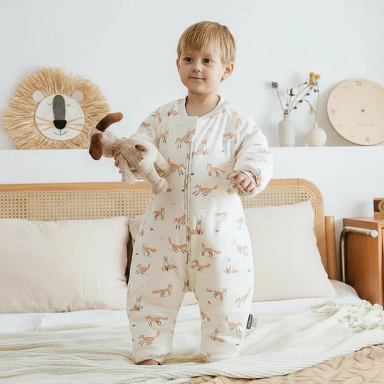Enfant portant une gigoteuse à jambes Kalinatur en coton bio, motif renard orange sur fond beige, debout sur un lit avec une peluche dans une chambre douce et naturelle.

