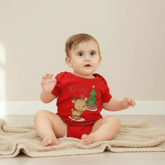 Bébé portant un body rouge Kalinatur motif renne et sapin de Noël, assis sur une couverture beige.
