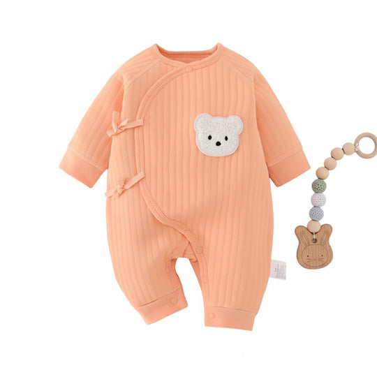 Combinaison bébé côtelée couleur pêche avec fermeture croisée et ourson brodé, présentée avec attache-sucette en bois.